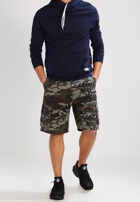 Merijakke med snøring, kengurulomme; kamuflasje cargo shorts med sidelommer; svarte joggesko med hvite detaljer.