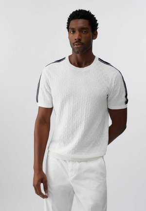 Bărbat în picioare cu expresie neutră, purtând o cămașă albă texturată cu mâneci scurte și dungi negre pe umeri și pantaloni albi, pe un fundal simplu.