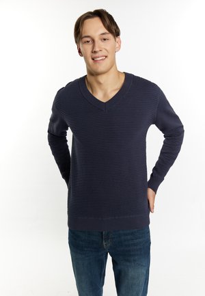 Marineblauwe v-hals trui met geribbelde textuur en lange mouwen, gedragen over een blauwe jeans. Model staat met handen op de heupen tegen een witte achtergrond.