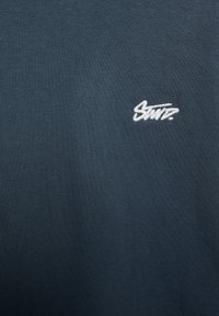 Mørkeblå, ribbet skjorte med et lille hvidt broderet logo på venstre side. Stoffet virker blødt og åndbart.