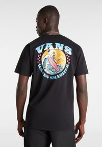 Černé bavlněné tričko s grafikou na zadní straně, zobrazující lebku surfující na růžovém prkně přes vlny a západ slunce, s textem "VANS WAVES SMASHER".