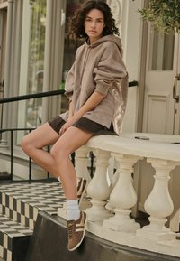 Brązowa, oversize'owa bluza wykonana z miękkiego materiału, zestawiona z ciemną spódnicą. Brązowe sneakersy z białymi paskami i skarpetkami. Ustawiona na stylowych schodach.