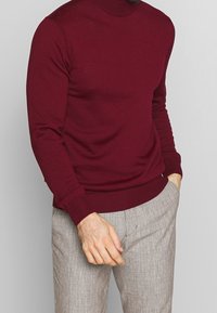 Burgunderfarbener gestrickter Pullover mit hohem Ausschnitt, schmaler Passform und gerippten Bündchen. Kombiniert mit grauen strukturierten Hosen.