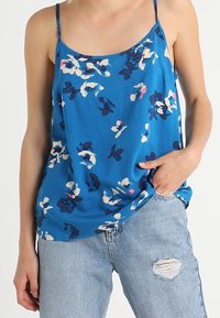 Persona che indossa un top senza maniche blu a fiori e jeans strappati blu chiaro, con una mano in tasca contro uno sfondo semplice.