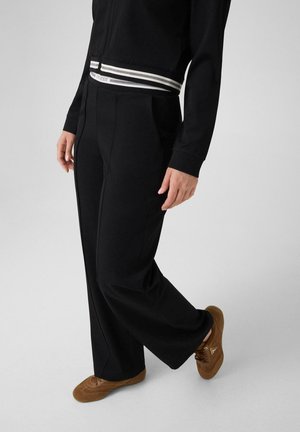 Personne portant un pantalon noir à jambes larges et une veste noire avec une ceinture rayée, associée à des baskets marron décontractées, debout sur un fond clair.