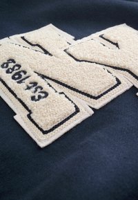 Marineblåt stof med beige, strukturerede lapper, der danner bogstaver og tal, omkranset af sort syning langs kanterne.