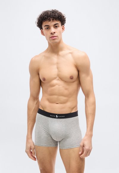 Άνδρας χωρίς πουκάμισο με κατσαρά μαλλιά που φοράει ανοιχτό γκρι μπόξερ με μαύρη μέση Ralph Lauren, στέκεται μπροστά σε ένα απλό φωτεινό φόντο.