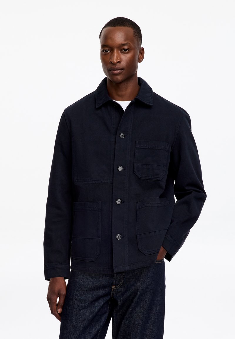 Homme portant une veste bleu marine foncé boutonnée avec trois poches devant et un jean foncé, debout devant un fond blanc uni.