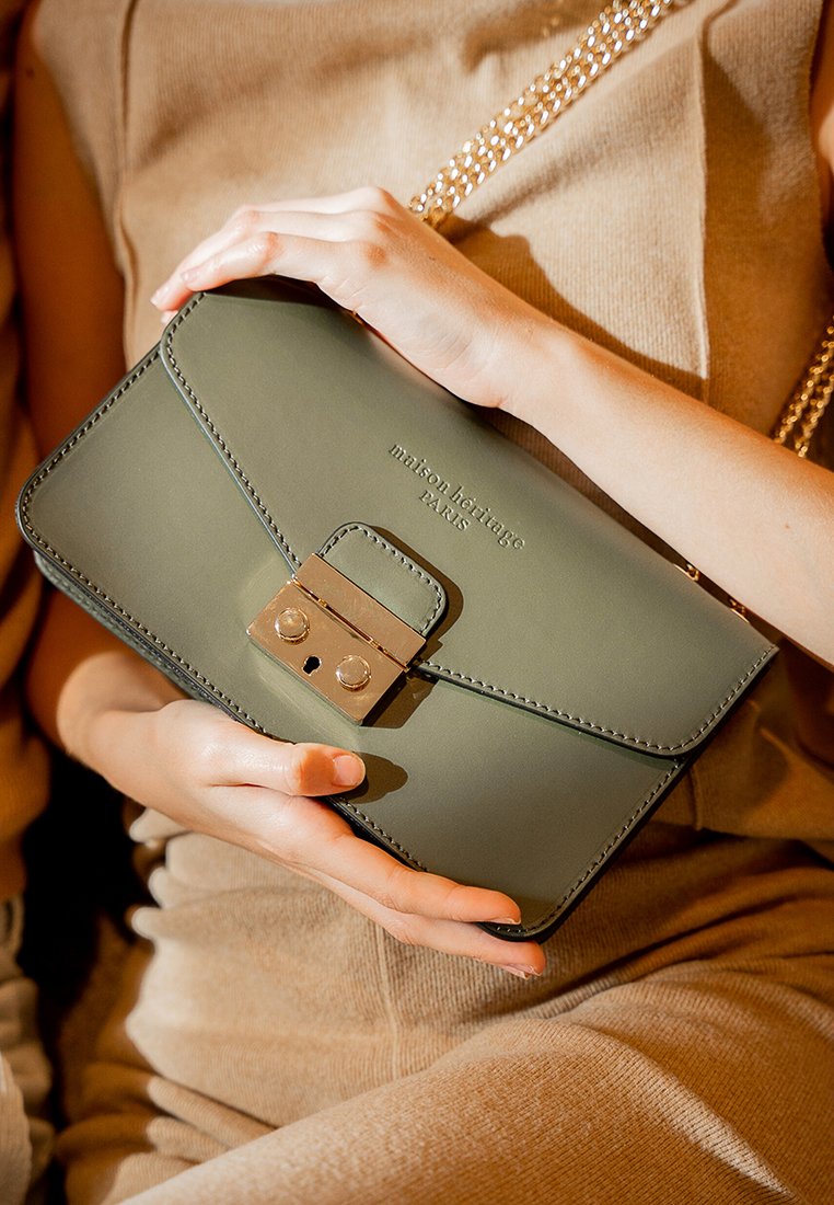 Clutch de cuero verde oliva con un cierre en tono dorado, que presenta el texto "maison heritage PARIS" en relieve en la parte frontal, sostenido por una mano.