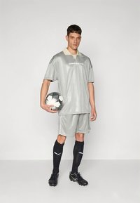 Άνδρας με γκρι αθλητική εμφάνιση Umbro FC κρατώντας μια ασπρόμαυρη ποδοσφαιρική μπάλα, φορά μαύρες κάλτσες Puma και παπούτσια ποδοσφαίρου, στέκεται μπροστά σε απλό φόντο.