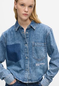 Πουκάμισο από τζιν με ανοιχτό μπλε απόχρωση, σκούρο μπλε τσέπη, κουμπιά μπροστά, δύο τσέπες στο στήθος και κεντημένο λογότυπο στην κάτω δεξιά πλευρά.