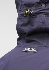 Navy waterproof jacket with hood, featuring a front zipper and drawstring, textured fabric, and a "Gaastra" label at the neck.