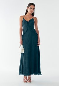 Abito teal con scollo all'americana, balze, tessuto testurizzato e una silhouette fluida. Abbinato a sandali con tacco bianchi e una piccola borsa bianca.