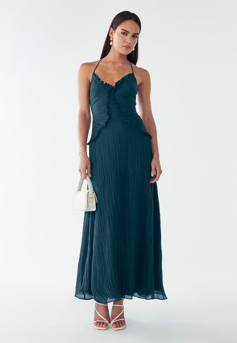 Abito teal con scollo all'americana, balze, tessuto testurizzato e una silhouette fluida. Abbinato a sandali con tacco bianchi e una piccola borsa bianca.