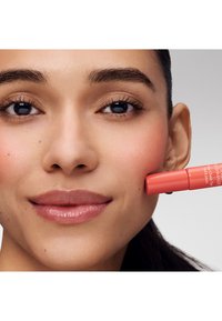 Kobieta nakłada koralowy róż na prawy policzek, prezentując gładką skórę, ciemne oczy i neutralny makijaż z subtelnym uśmiechem.