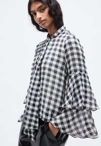 Μπλούζα με ασπρόμαυρο καρό σχέδιο και μανίκια με βολάν τύπου καμπάνας, κατασκευασμένη από ελαφρύ ύφασμα. Διαθέτει γιακά και κλείσιμο με κουμπιά μπροστά.