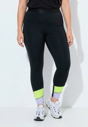 Leggings mit schwarzem Oberteil, neongelben und hellvioletten Knöchelriemen, getragen mit weißen Sportschuhen, stehend an einer weißen Wand.