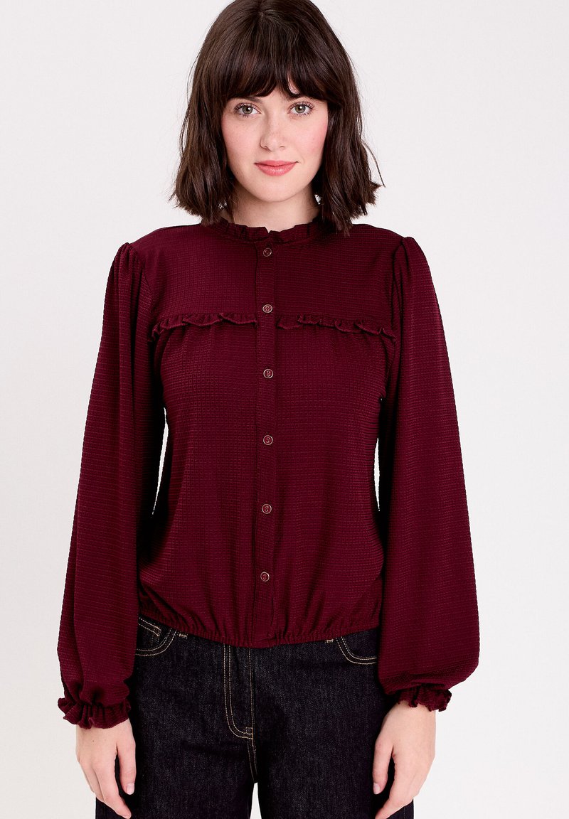 Burgunderfarbene Bluse mit Knopfleiste und Rüschendetails an der Brust und den Manschetten, aus strukturiertem Stoff gefertigt, mit einem elastischen Saum in der Taille.