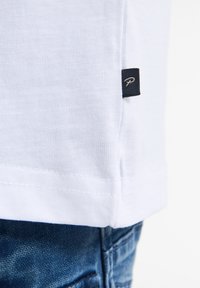 Camiseta blanca de manga corta hecha de algodón, con una pequeña etiqueta negra que lleva un logo en el dobladillo, combinada con pantalones de mezclilla azules.