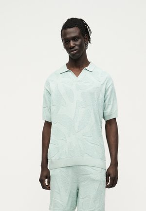 Tânăr cu piele închisă la culoare și dreadlocks, purtând un tricou polo texturat cu mânecă scurtă bleu și pantaloni scurți asortați, pe un fundal simplu.