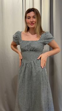 Moteris su peties ilgio šviesiais plaukais, dėvinti juodai baltą languotą suknelę su pūstomis rankovėmis, stovinti su rankomis ant klubų priešais užuolaidą.