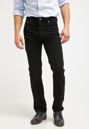 Mand iført sorte slim-fit jeans, lyseblå skjorte med knapper og op rullede ærmer, samt sorte lædersko, stående.