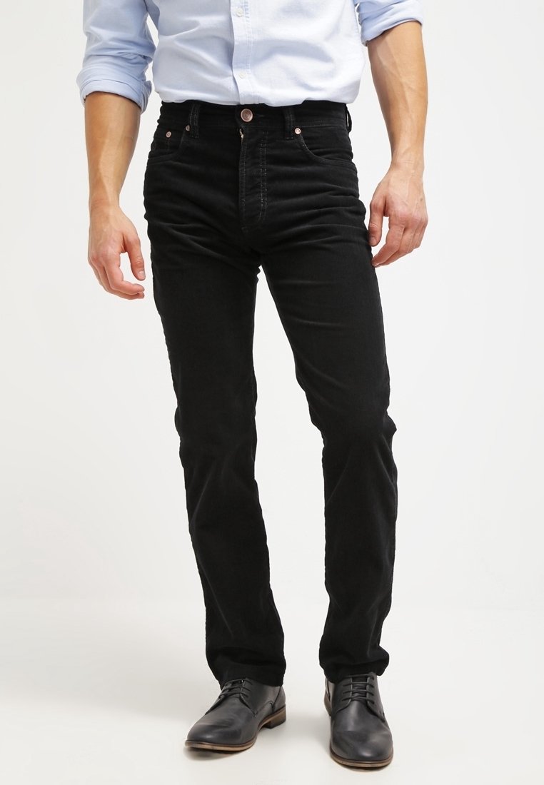 Homme portant un jean slim noir, une chemise bleu clair boutonnée avec manches retroussées, et des chaussures habillées en cuir noir, debout.