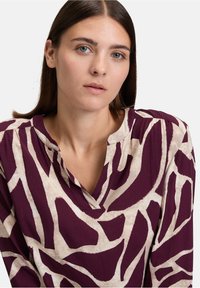 Blusa a maniche lunghe di un profondo viola con un motivo astratto beige. Presenta un collo con apertura parziale e una texture di tessuto morbido e liscio.