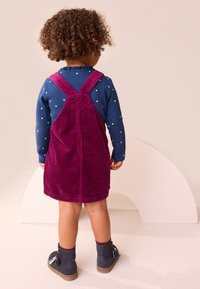 Bambino che indossa un vestito salopette in velluto a coste bordeaux sopra una maglia blu a maniche lunghe con pois bianchi, abbinato a stivali scuri.