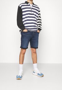 Randig långärmad tröja i marinblått och vitt med svarta ärmar, tillsammans med mörka denimshorts. Blå och vita sneakers fulländar looken.