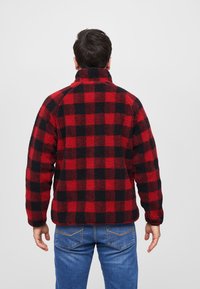 Rød og sort ternet fleecejakke med høj krave, struktureret overflade og en afslappet pasform, parret med blå denimjeans.