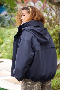 Persoană cu părul creț, îmbrăcată într-o jachetă cu glugă de culoare bleumarin și pantaloni cu model de camuflaj, stând în aer liber lângă vegetation și o barcă mică.