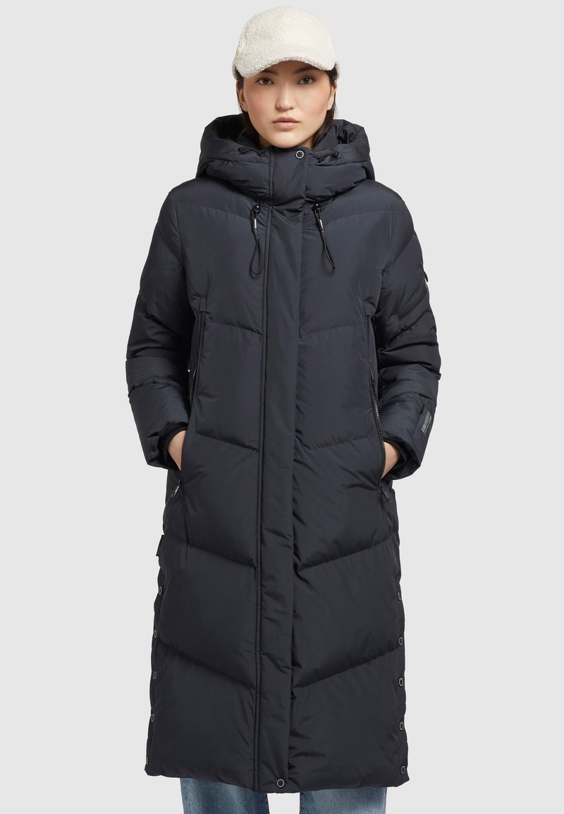 Donna che indossa un lungo cappotto invernale imbottito nero con cappuccio, mani nelle tasche laterali e un berretto bianco testurizzato, in piedi contro uno sfondo semplice.