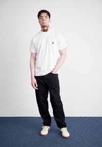 Mladý muž s vitiligo, oblečený v bílém tričku, černých kalhotách a bílých teniskách, stojící proti bílé zdi na modrém koberci.