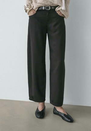 Donna con pantaloni neri a gamba larga, cintura nera, maglione beige in maglia e ballerine nere, in piedi con le mani nelle tasche.