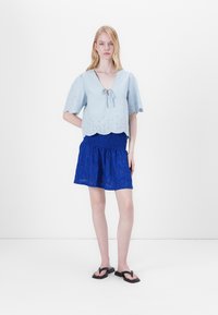 Ανοιχτό μπλε κομμένο μπλουζάκι με κροσέ στρίφωμα και λεπτομέρειες από ματάκια, συνδυασμένο με μια υφασμάτινη μπλε φούστα. Το μοντέλο φορά μαύρες σαγιονάρες.