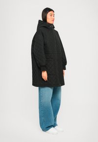 Vrouw in een oversized zwarte gewatteerde jas met capuchon, brede blauwe jeans en witte sneakers, staande tegen een effen achtergrond.