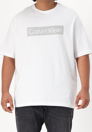 Osoba ubrana w białą koszulkę Calvin Klein z szarym paskowanym logo i czarne spodnie, pokazana od szyi do bioder.
