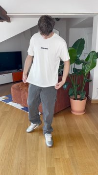 Bijela pamučna majica s crnim natpisom "SHINJUKU" na prsima, u kombinaciji s velikim sivoj trapericama i srebrnim sportskim cipelama, u kućnom okruženju.