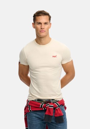 Uomo che indossa una maglietta beige aderente con logo rosso, jeans blu e un maglione a righe rosse legato intorno alla vita, in piedi con le mani dietro la schiena.