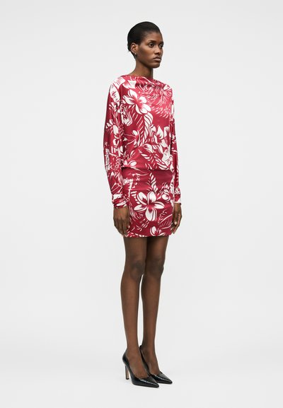 Γυναίκα που στέκεται φορώντας κόκκινη και λευκή φλοράλ μπλούζα με μακριά μανίκια και κοντή φούστα που ταιριάζει, μαζί με μαύρα μυτερά ψηλά τακούνια.