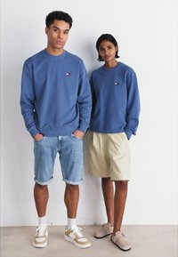 Sudaderas de cuello redondo azules en modelos masculinos y femeninos, combinadas con pantalones cortos de mezclilla y pantalones cortos ligeros, respectivamente. Logotipo destacado en las sudaderas.