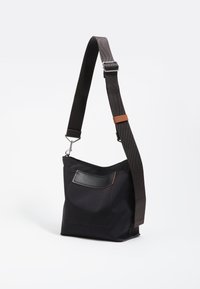 Bolsa de tela negra con una correa para el hombro, que presenta un bolsillo frontal con un acento de cuero y detalles de costura en marrón. Diseño compacto y moderno.
