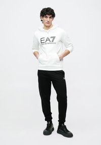 Felpa con cappuccio bianca con logo nero "EA7 Emporio Armani", abbinata a pantaloni neri. Entrambi gli articoli sono realizzati in tessuto liscio, con una vestibilità comoda.
