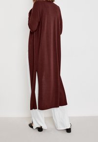 En person iförd en lång vinröd cardigan med sidoklyftor, vita vida byxor och svarta öppna klacksandaler, stående mot en vit vägg.