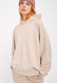 Beige hooded sweatshirt with a soft texture and ribbed cuffs. Features a small embroidered logo on the front. Worn with light-colored pants.