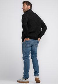 Zwarte gebreide trui met een hoge kraag, gecombineerd met lichtblauwe spijkerbroek. Het model draagt grijze sneakers met bruine zolen.