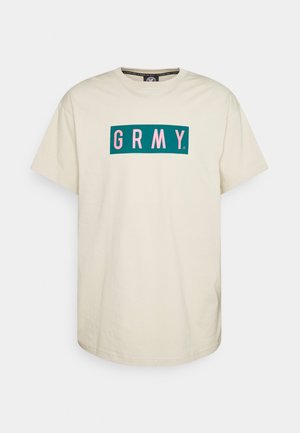 Beżowy T-shirt z krótkim rękawem z zielonym prostokątnym logo na piersi, na którym widnieją różowe litery "GRMY".