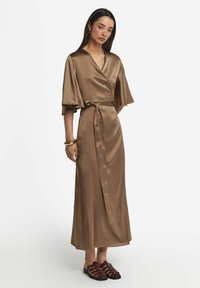 Braunes Satin-Wickelkleid mit tiefem V-Ausschnitt, kurzen Ärmeln und Gürtelakzent. Model trägt braune Riemchensandalen. Schlichtes Hintergrund.
