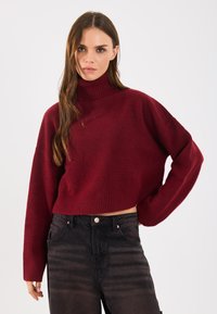 Burgunderfarbener Cropped-Pullover mit hohem, geripptem Rollkragen, der eine weiche Textur und weite Ärmel aufweist, kombiniert mit hochgeschnittenen schwarzen Jeans.